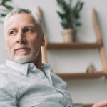 A older white man sits looking out a window with a tablet in his hand, acting as a financial advisor and counselor