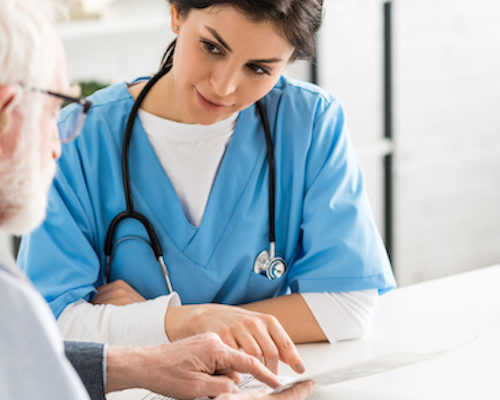 A woman wearing scrubs and a stethoscope helps an older man with paperwork