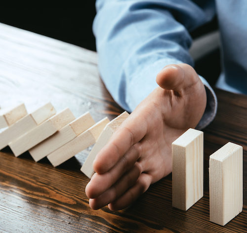 A man sits at a table with blocks in a line. His hand intersects a line of block stopping the flow of blocks cascading, implying sequence risk protection.